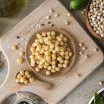 Bol de pois chiches cuits posé sur une planche en bois, entouré de pois chiches secs, de sel et d’assaisonnements.