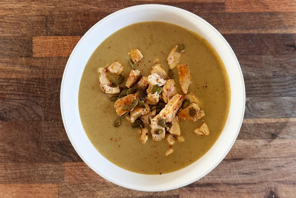 Velouté de fèves servi dans un bol, garni de morceaux de poulet dorés et de graines de courge, posé sur une table en bois avec une cuillère à côté.