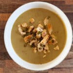 Velouté de fèves servi dans un bol, garni de morceaux de poulet dorés et de graines de courge, posé sur une table en bois avec une cuillère à côté.