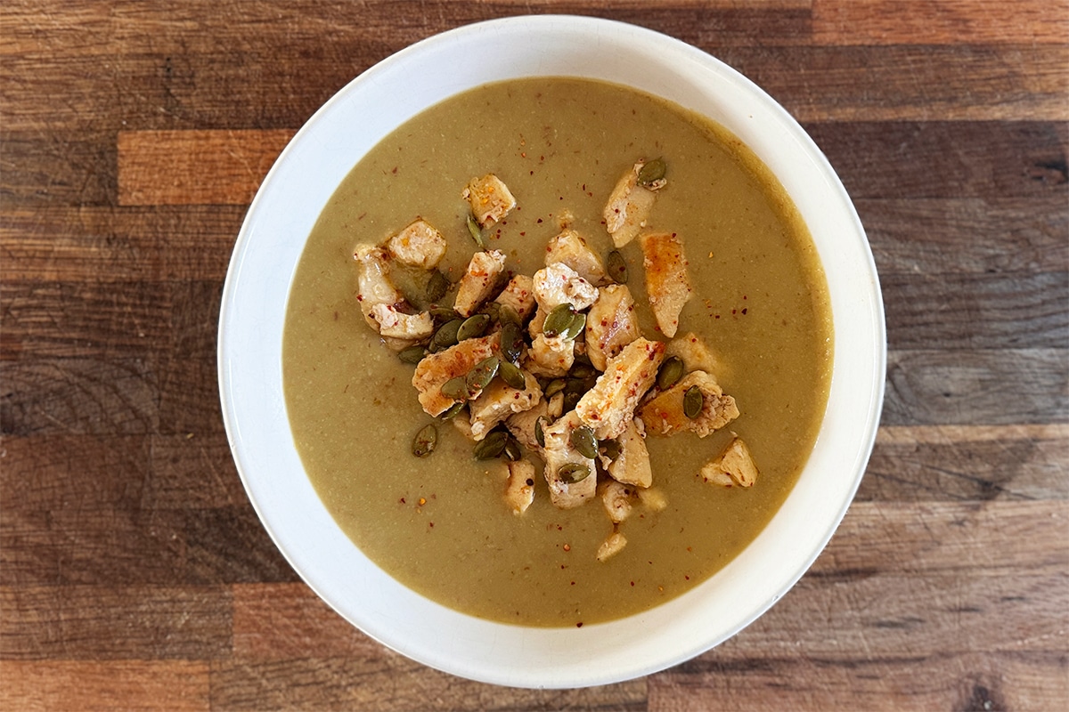 Velouté de fèves servi dans un bol, garni de morceaux de poulet dorés et de graines de courge, posé sur une table en bois avec une cuillère à côté.