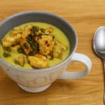 Velouté de fèves servi dans un bol, garni de morceaux de poulet dorés et de graines de courge, posé sur une table en bois avec une cuillère à côté.